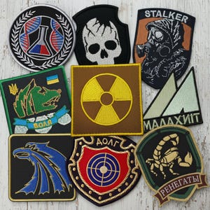 May include: A collection of embroidered patches with various designs. Included are patches with a skull, a gas mask, a radiation symbol, a scorpion, and a blue eagle. The patches are in various shapes and colors, including black, brown, green, and blue.