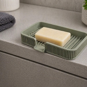 May include: A sage green soap dish holding a bar of soap. The dish is rectangular with vertical lines on the sides and a slatted base. A grey towel, a white ceramic container, and a potted plant are in the background.