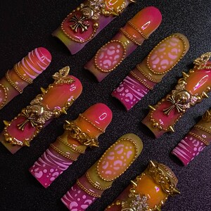 May include: A collection of vibrant, ornate press-on nails. Each nail features a gradient of pink, orange, and yellow, adorned with gold-colored embellishments, including chains, studs, and decorative charms. Some nails have leopard print or striped designs.