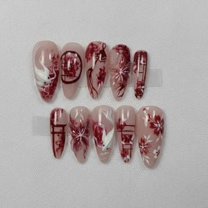 May include: A set of almond-shaped false nails with a nude base, featuring intricate designs in red and white. The designs include floral patterns, stylized dragons, and architectural elements, creating an artistic and elegant aesthetic.