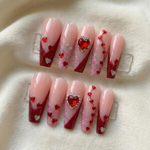 May include: Set of ten artificial nails with a light pink base, red glitter accents, and heart-shaped embellishments. Some nails feature a large, heart-shaped jewel. Silver and red hearts are scattered across the nails.