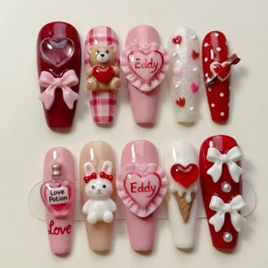 May include: A set of ten artificial nails with various designs. The nails feature hearts, bows, teddy bears, and the word "Love". Colors include red, pink, white, and beige. Some nails have the name "Eddy" on them.