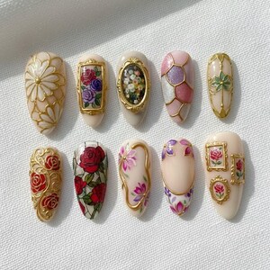May include: A collection of ten almond-shaped artificial nails with intricate designs. The nails feature floral patterns, geometric shapes, and framed miniature paintings in gold, red, pink, purple, and white. The designs are detailed and elegant.