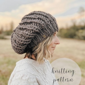 May include: A brown knitted hat with a ribbed design. The hat has a tag with initials. The image also includes the text "knitting pattern". The hat is worn by a person wearing a light-colored sweater.
