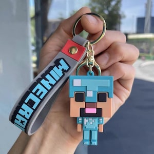May include: A Minecraft-themed keychain featuring a pixelated character in shades of blue, tan, and black. The keychain includes a gray strap with the word "Minecraft" in white letters and a gold-colored keyring.