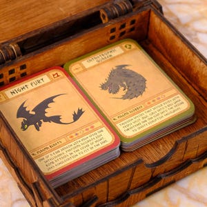 May include: Wooden box with a hinged lid, containing a stack of trading cards. The cards feature illustrations of dragons, with text including "Night Fury" and "Catastrophic Quereen". The box is made of light brown wood.