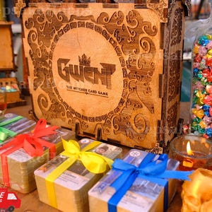 May include: Wooden box with the text "Gwent: The Witcher Card Game" and decorative carvings. Several card decks wrapped with colorful ribbons are in front of the box. A jar filled with colorful origami stars is also visible.