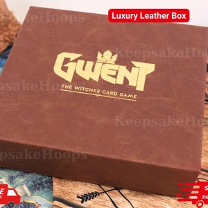 May include: A brown leather box with the text "Gwent The Witcher Card Game" in gold lettering. The box has a textured surface and the words "Luxury Leather Box" are in a red box in the top right corner.