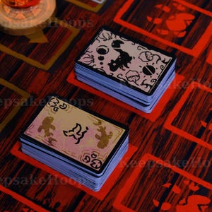 May include: Two stacks of illustrated cards with black and white designs sit on a red and brown patterned surface. The cards have a light beige border and feature various whimsical illustrations. The background has red and yellow geometric shapes.