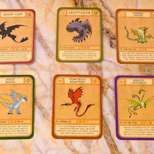 Puede incluir: Seis cartas coleccionables con ilustraciones de dragones. Cada carta presenta un diseño de dragón único con un nombre, descripción y estrellas de calificación. Las cartas tienen un fondo beige con bordes de colores: rojo, verde, marrón y morado.