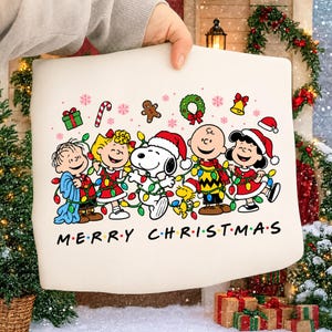 Może przedstawiać: Kremowa bluza z grafiką przedstawiającą postacie z Fistaszków, w tym Snoopy'ego i Charliego Browna, noszące czapki Mikołaja i zaplątane w kolorowe lampki choinkowe, z nadrukowanym poniżej napisem MERRY CHRISTMAS.
