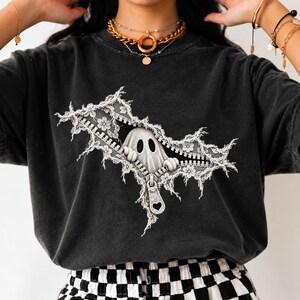 May include: A charcoal gray short-sleeve t-shirt featuring a graphic print of a white ghost peeking through a zipper opening. The zipper is surrounded by white lace-like floral patterns and includes a heart-shaped pull tab detail.