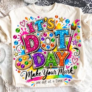 Op de afbeelding: Een crèmekleurig t-shirt met een kleurrijke afbeelding met de tekst IT'S DOT DAY en Make Your Mark one dot at a time. Het ontwerp bevat glinsterende letters, een verfpalet, een penseel en diverse kleine stippen en sterren.