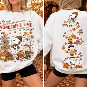 Puede incluir: Una sudadera blanca de manga larga con personajes de Peanuts en un entorno otoñal. El frente muestra a Snoopy y Charlie Brown con el texto The most WONDERFUL TIME of the year. La parte posterior muestra una enredadera vertical de hojas de otoño con Snoopy, Charlie Brown, Woodstock y Lucy.