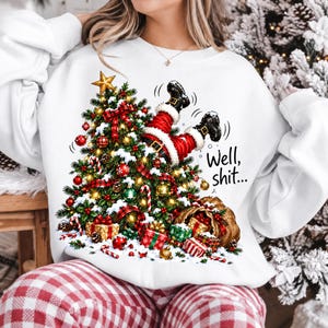 May include: A white crewneck sweatshirt featuring a graphic of a decorated Christmas tree with a pair of Santa Claus legs sticking out of the top. The text "Well, shit..." is printed in black script next to the tree, which is surrounded by gifts.