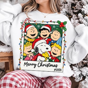Może przedstawiać: Biała bluza z długim rękawem z grafiką postaci z Peanuts w ramce w stylu polaroidu, ozdobiona świątecznymi światełkami, ostrokrzewem i laską cukrową. Napis Merry Christmas oraz #2026 znajduje się pod postaciami wewnątrz ramki.