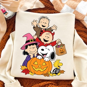 May include: A cream-colored sweatshirt featuring a colorful Peanuts Halloween graphic. The design shows Linus, Charlie Brown in a devil costume, Lucy in a witch hat, Snoopy behind a jack-o'-lantern, and Woodstock in a skeleton costume with autumn leaves.