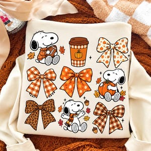 Könnte beinhalten: Ein cremefarbenes Sweatshirt mit einem Grafikdruck von Snoopy in verschiedenen herbstlichen Posen, einem Kaffeebecher mit Kürbismotiv sowie mehreren dekorativen Schleifen in Karomustern, Leopardenmuster und Blattmotiven in Orange.