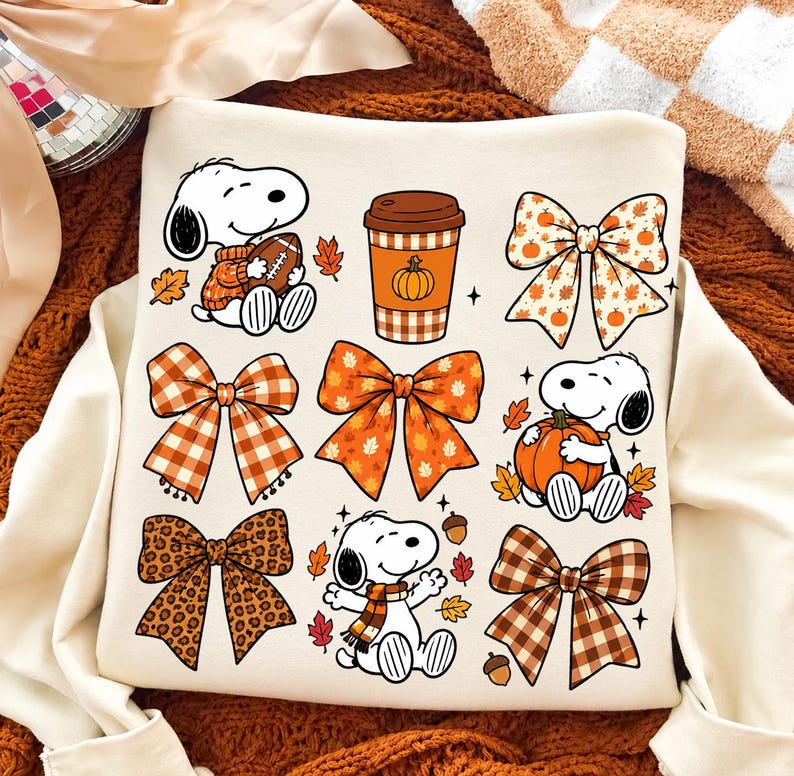 Może przedstawiać: Kremowa bluza z grafiką przedstawiającą Snoopy'ego w r&oacute;żnych jesiennych pozach, kubek do kawy z motywem dyni oraz kilka ozdobnych kokard w kratę, panterkę i wzory liści w odcieniach pomarańczu i brązu.