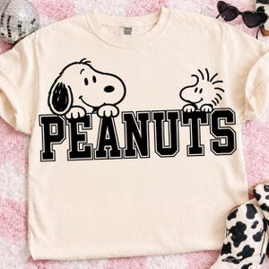 Może przedstawiać: Kremowy t-shirt z krótkim rękawem z czarną grafiką przedstawiającą Snoopy'ego i Woodstocka wyglądających znad słowa PEANUTS napisanego pogrubioną czcionką blokową. Koszulka ma okrągły dekolt i widoczną metkę marki wewnątrz kołnierzyka.