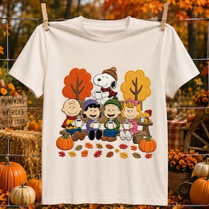 May include: A cream-colored short-sleeve t-shirt featuring a graphic print of the Peanuts characters Snoopy, Charlie Brown, Lucy, Linus, Sally, and Woodstock sitting on a bench holding mugs, surrounded by autumn trees, pumpkins, and leaves.