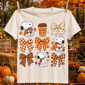 Może przedstawiać: Kremowy t-shirt z kr&oacute;tkim rękawem z siatką ilustracji przedstawiających Snoopy'ego trzymającego piłkę do futbolu, kubek kawy z dynią, Snoopy'ego z dynią, Snoopy'ego w szaliku oraz r&oacute;żne wzorzyste kokardy w kolorach pomarańczowym i brązowym.