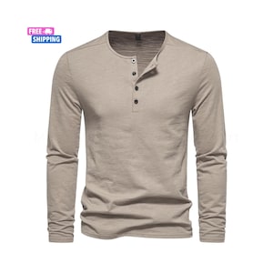 May include: A long-sleeved, light brown Henley shirt with a crew neck and three black buttons. The shirt is made of a soft, textured fabric and has a classic, casual design. The shirt is a neutral color.