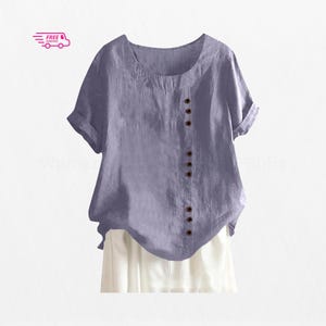 May include: A short-sleeved, lavender-coloured shirt with a round neckline and a vertical row of dark brown buttons offset to the right side. The sleeves feature a folded cuff detail, and the fabric has a visible, slightly textured weave.