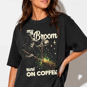 Op de afbeelding: Een zwart t-shirt met korte mouwen en een afbeelding van een skeletheks die koffie op een bezemsteel giet. De tekst "my Broom runs ON COFFEE" staat in een crèmekleurig lettertype. Het ontwerp bevat kleine steraccenten en groene vonken.