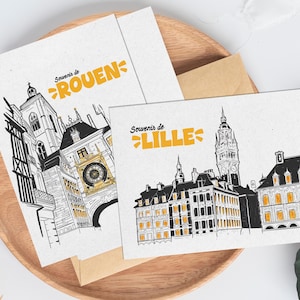 May include: Two postcards with architectural illustrations of French cities, Rouen and Lille, in black and white with yellow accents. The postcards are on a wooden tray with a brown envelope.