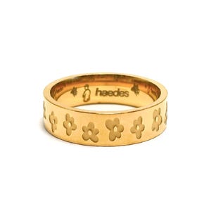 Op de afbeelding: Goudkleurige ring met een herhalend bloemmotief rond de band. De ring is gegraveerd met het woord "heedes" en een kleine illustratie. De ring is een eenvoudige bandring.