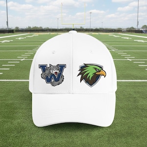 May include: White baseball cap featuring two embroidered logos. One logo depicts a snarling wolf over a blue "W", the other shows a green and brown eagle head within a shield. The cap is set against a green football field.