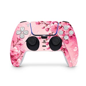 Könnte beinhalten: Ein rosa-weißes PlayStation 5 Controller-Skin mit einem floralen Kirschblüten-Design. Der Controller hat schwarze Joysticks und Knöpfe, mit einem hellrosa Hintergrund und einem Muster aus rosa Blüten.