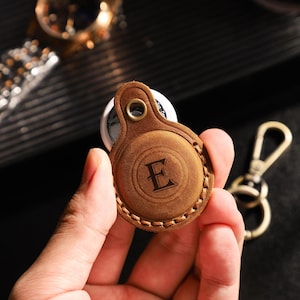 Könnte beinhalten: Ein brauner Leder-AirTag-Halter mit dem Buchstaben "E" auf der Vorderseite. Der Halter hat einen genähten Rand und einen goldfarbenen Metall-Schlüsselring. Der Hintergrund zeigt eine Uhr und eine dunkle Oberfläche.