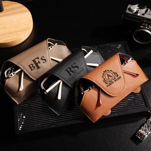 May include: Three personalized eyeglass cases in brown, black, and tan leather, each with a pair of glasses inside. The cases have embossed initials or a name. A watch, camera, and Eiffel Tower figurine are in the background.