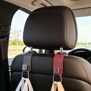 May include: Car headrest hooks in gray and burgundy leather. Each hook has a silver metal ring for hanging bags. The hooks are attached to the headrest posts and are designed to keep bags off the car floor.