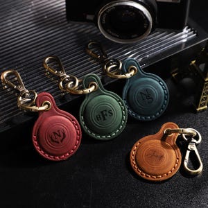 May include: A collection of personalized leather keychains in various colors: red, green, teal, and brown. Each keychain features a gold-colored clasp and is embossed with initials. The keychains are circular and have a stitched border.
