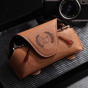 Puede incluir: Un estuche de gafas de cuero marrón con el texto "Cherish Best" y un diseño de corona de laurel. El estuche está abierto, revelando un par de gafas con montura marrón. Una cámara negra está en el fondo.