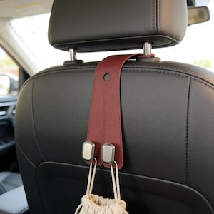 May include: A burgundy leather car seat headrest organizer with two silver hooks. A beige drawstring bag hangs from the hooks. The organizer is attached to a black car seat.