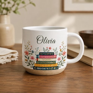 Puede incluir: Taza de cerámica blanca con el nombre "Olivia" en cursiva. La taza presenta una pila de libros coloridos y detalles florales. La base de la taza tiene un acabado natural sin esmaltar. Un regalo perfecto para los amantes de los libros.