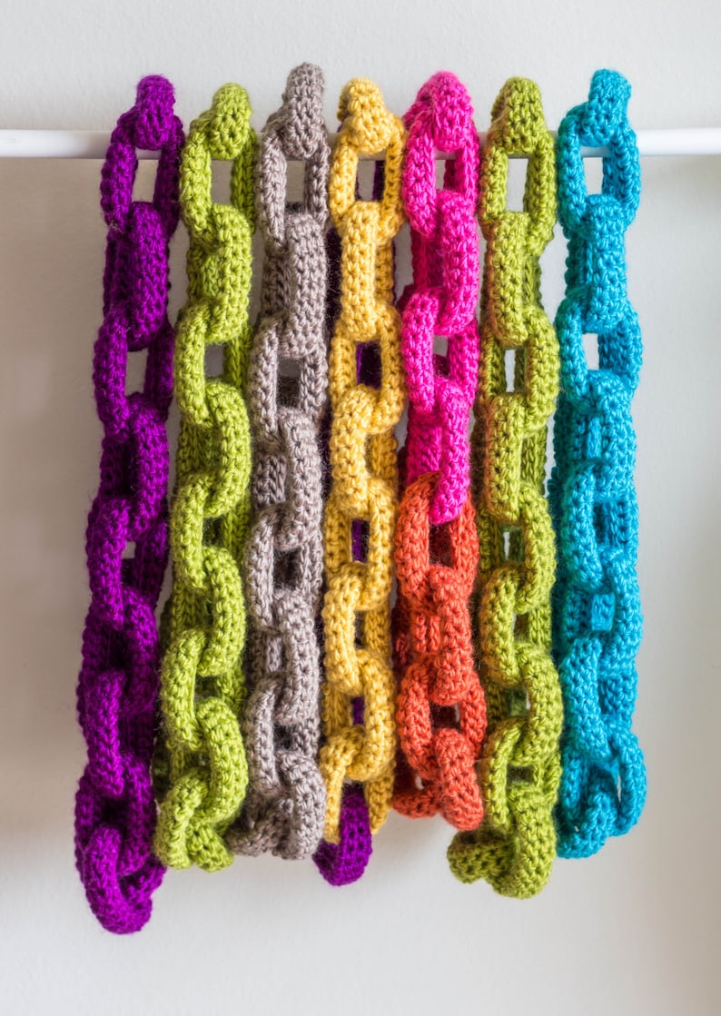 May include: Seven crocheted chains in different colours hanging from a white bar. The chains are purple, green, grey, yellow, orange, red, and blue.