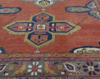 May include: A detailed shot of a red area rug with an intricate geometric pattern. The rug features a central design with blue, yellow, and pink accents. The border has a repeating floral and bird motif in gray, pink, and yellow. The rug has a fringed edge.