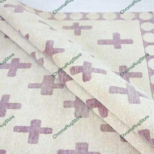Può includere: Un tappeto avvolto, bianco sporco, con un motivo ripetuto di croci lavanda. Il tappeto ha un bordo di forme geometriche lavanda e un bordo sfrangiato. La trama del tappeto è visibile, suggerendo un materiale tessuto.