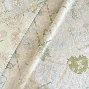 Op de afbeelding: Close-up van een crèmekleurig tapijt met een bloemenpatroon in tinten groen, blauw en beige. Het tapijt heeft een distressed, vintage look met zichtbare vouwen en texturen. Het ontwerp bevat gestileerde bloemen en geometrische vormen.