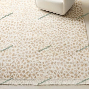 May include: A beige and white patterned rug with a leopard print design. The rug has a fringed edge and is partially visible with a white chair leg in the upper right corner. The rug is made of a soft material.