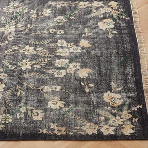 May include: A distressed black area rug with a floral pattern in cream and beige. The rug has a vintage look with a fringed edge. The floral design includes flowers and leaves.