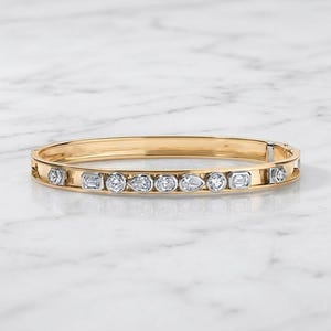 Könnte beinhalten: Ein goldener Armreif mit einer Reihe abwechselnder, klarer Edelsteine in verschiedenen Formen, darunter runde, tropfenförmige und rechteckige Schliffe, die in silberfarbenen Fassungen auf der Vorderseite des polierten Goldbandes sitzen.