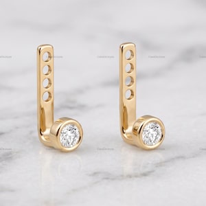 May include: A pair of gold ear jackets featuring a vertical bar with four circular cutouts and a single round brilliant-cut diamond set in a bezel at the base. The jewelry pieces are polished and have a smooth, reflective metallic finish.