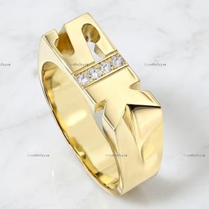 Op de afbeelding: Een gouden ring met de letters S en K als centraal designelement. De letter K is versierd met drie kleine, heldere, rondgeslepen stenen in een horizontale rij. De ring heeft een gepolijste, glanzende afwerking en een opvallende vorm.