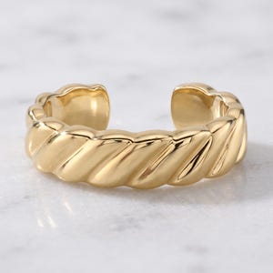 May include: A gold-toned open ring featuring a continuous twisted rope texture along the entire band. The metal has a polished, reflective finish with a distinct gap at the back, allowing for slight adjustment in the fit of the circular band.